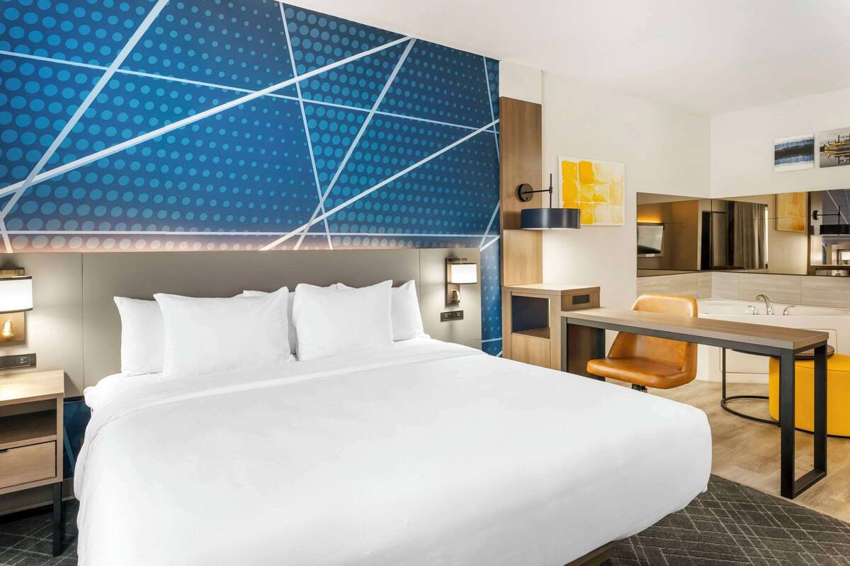 A hotel room with a large bed, a desk, a chair, a small table, a sink, and a mirror.