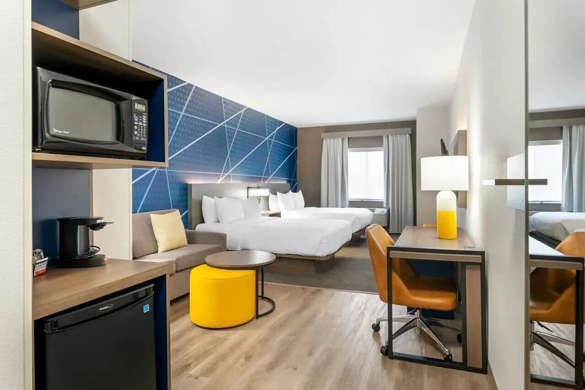 A hotel room with a bed, a desk with a microwave, a coffee maker, a yellow ottoman, and a chair.
