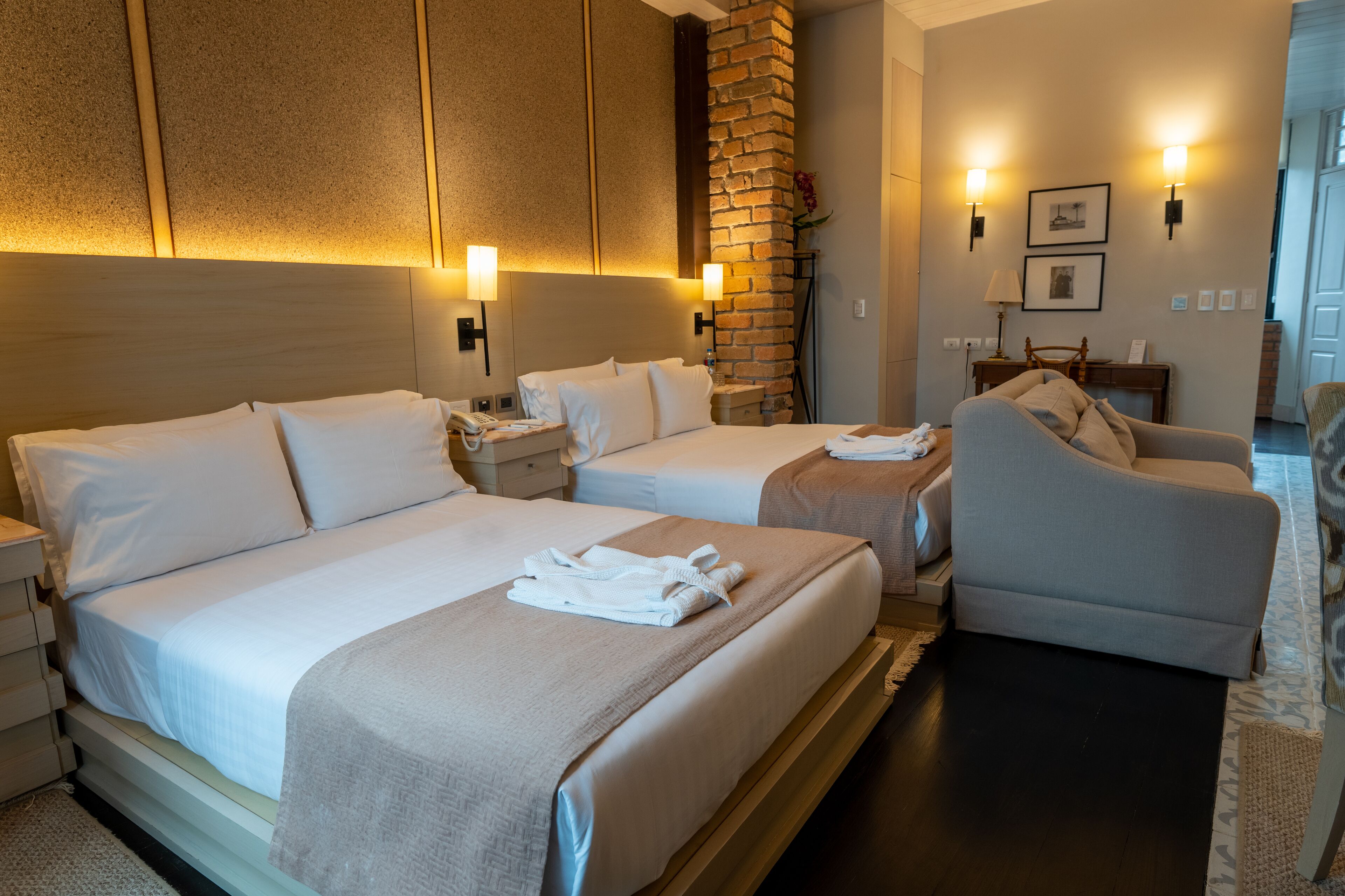 Habitación de hotel con dos camas, una zona de estar con una silla, una mesita y una pared de ladrillo decorativa.