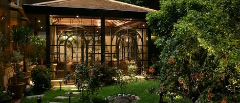 Une véranda dotée d’un toit en verre, entourée d’un jardin où poussent différentes espèces de plantes, et agrémentée d’un banc.