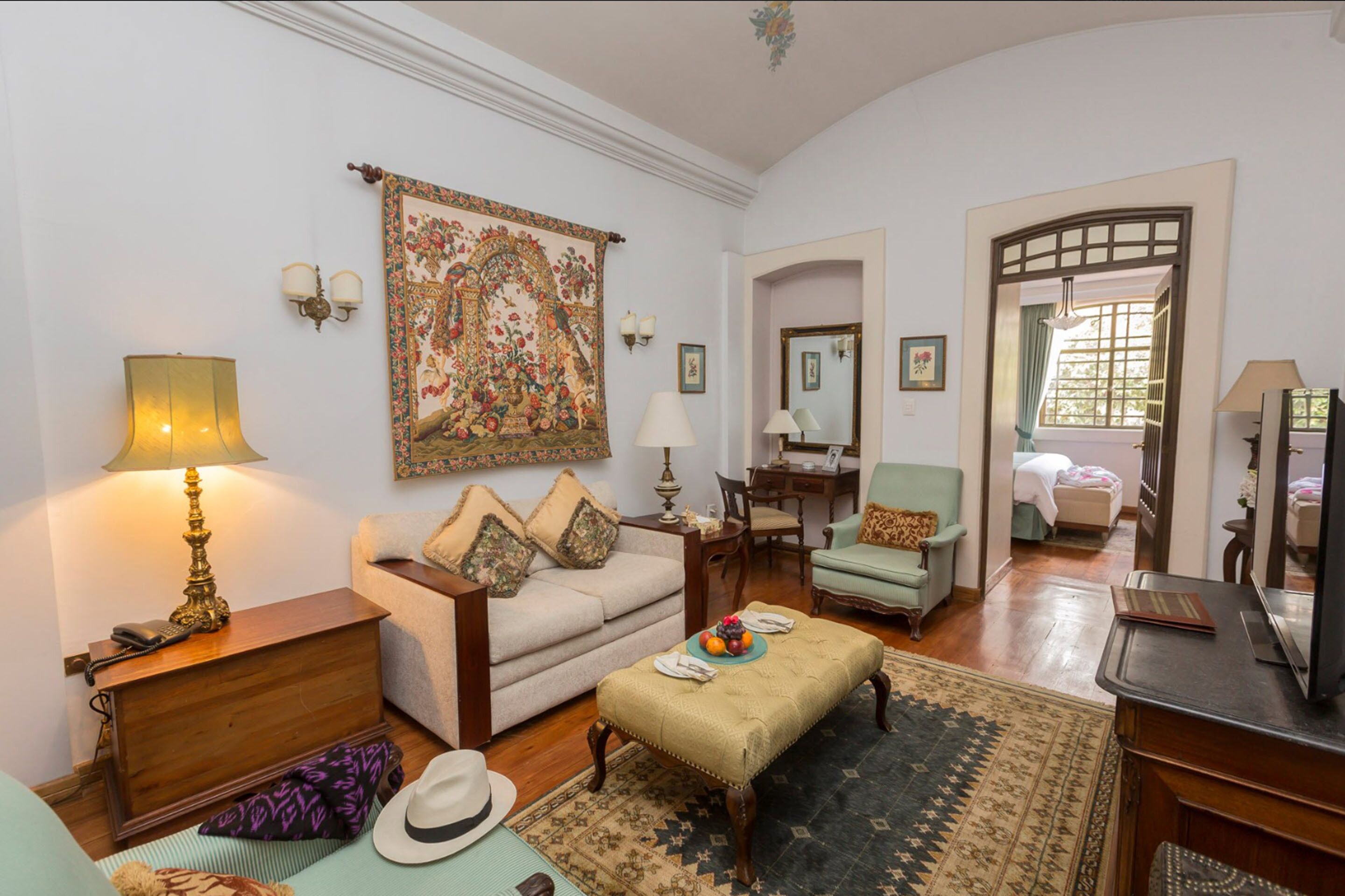 Un salón tradicional con sofá, sillones, una mesa de centro y una mesa de comedor. Hay un tapiz grande en la pared, un espejo y una ventana con cortinas.