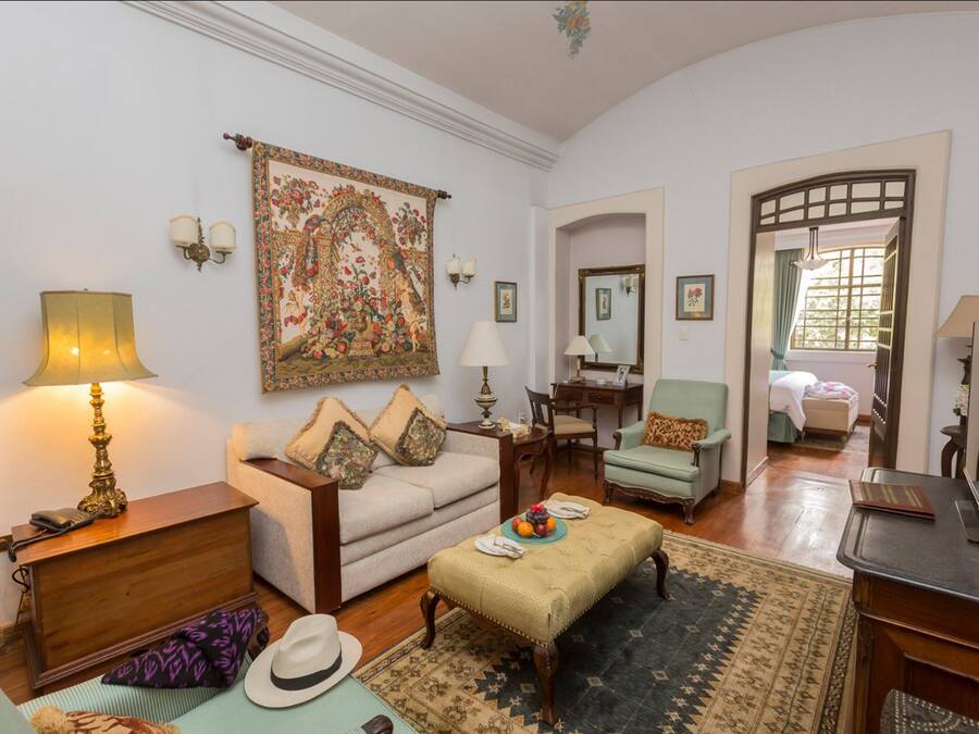 A traditional living room with a sofa, armchairs, a coffee table, and a dining table. There is a large tapestry on the wall, a mirror, and a window with curtains.