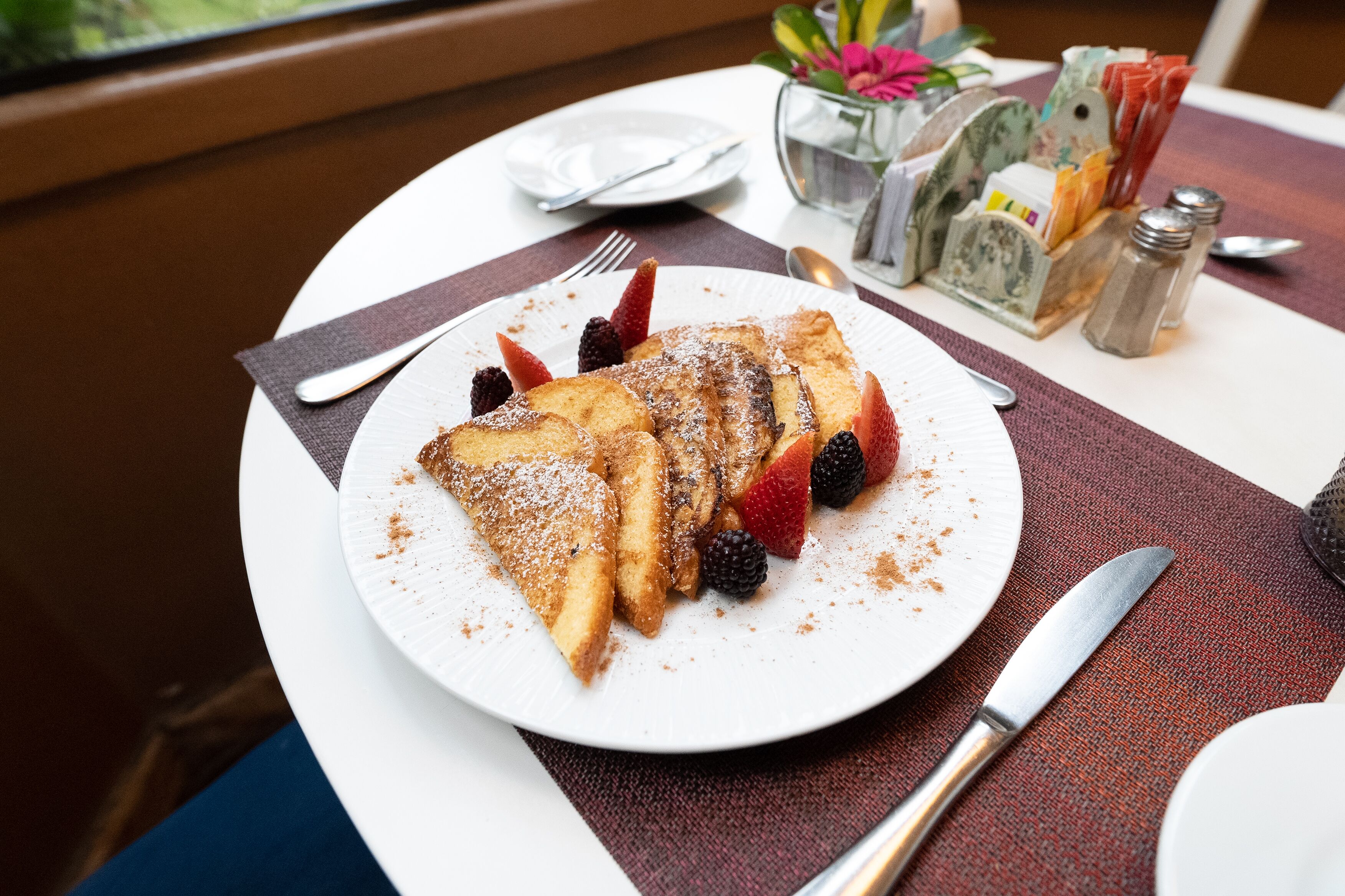A breakfast plate with pancakes, strawberries, and blackberries, a knife, and a spoon on a table with a placemat.