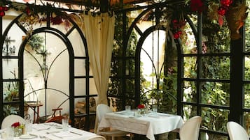 Ein Restaurant mit gedeckten Tischen, umgeben von großen Fenstern und dekorativen Pflanzen und Blumen.