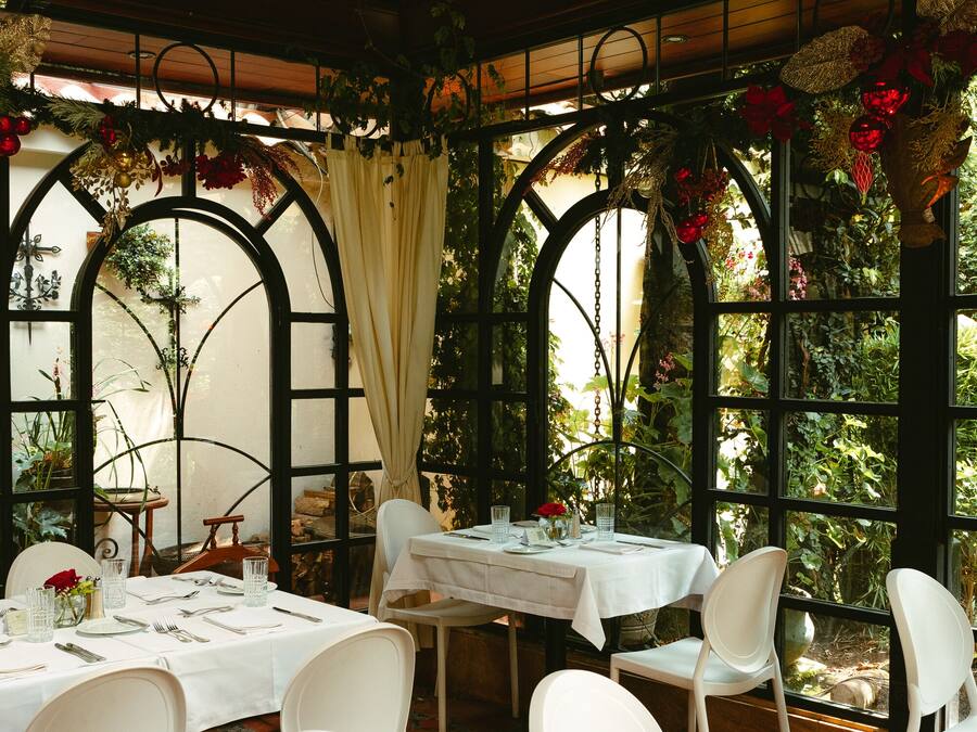 Ein Restaurant mit gedeckten Tischen, umgeben von großen Fenstern und dekorativen Pflanzen und Blumen.