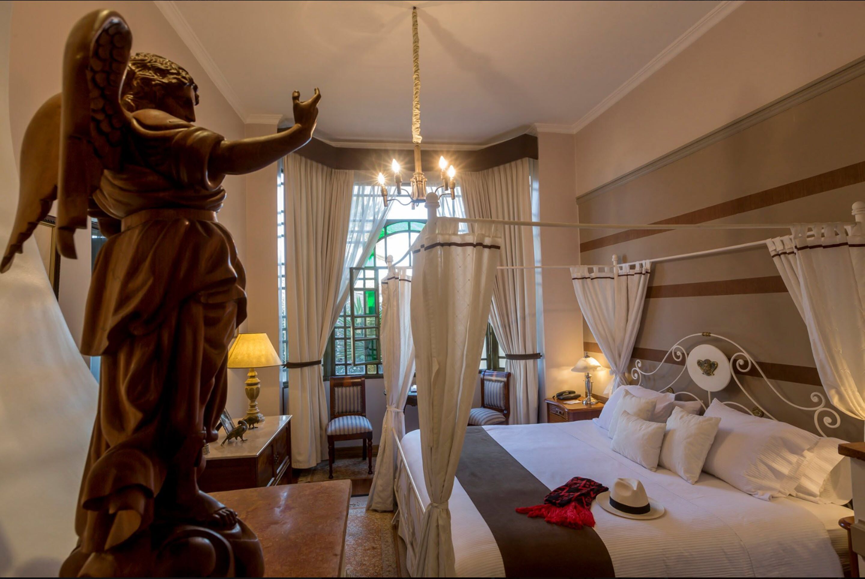 Ein Hotelzimmer mit einer großen Engelsstatue, einem Himmelbett, einem Chandelier und Blick auf einen grünen Garten durch das Fenster.
