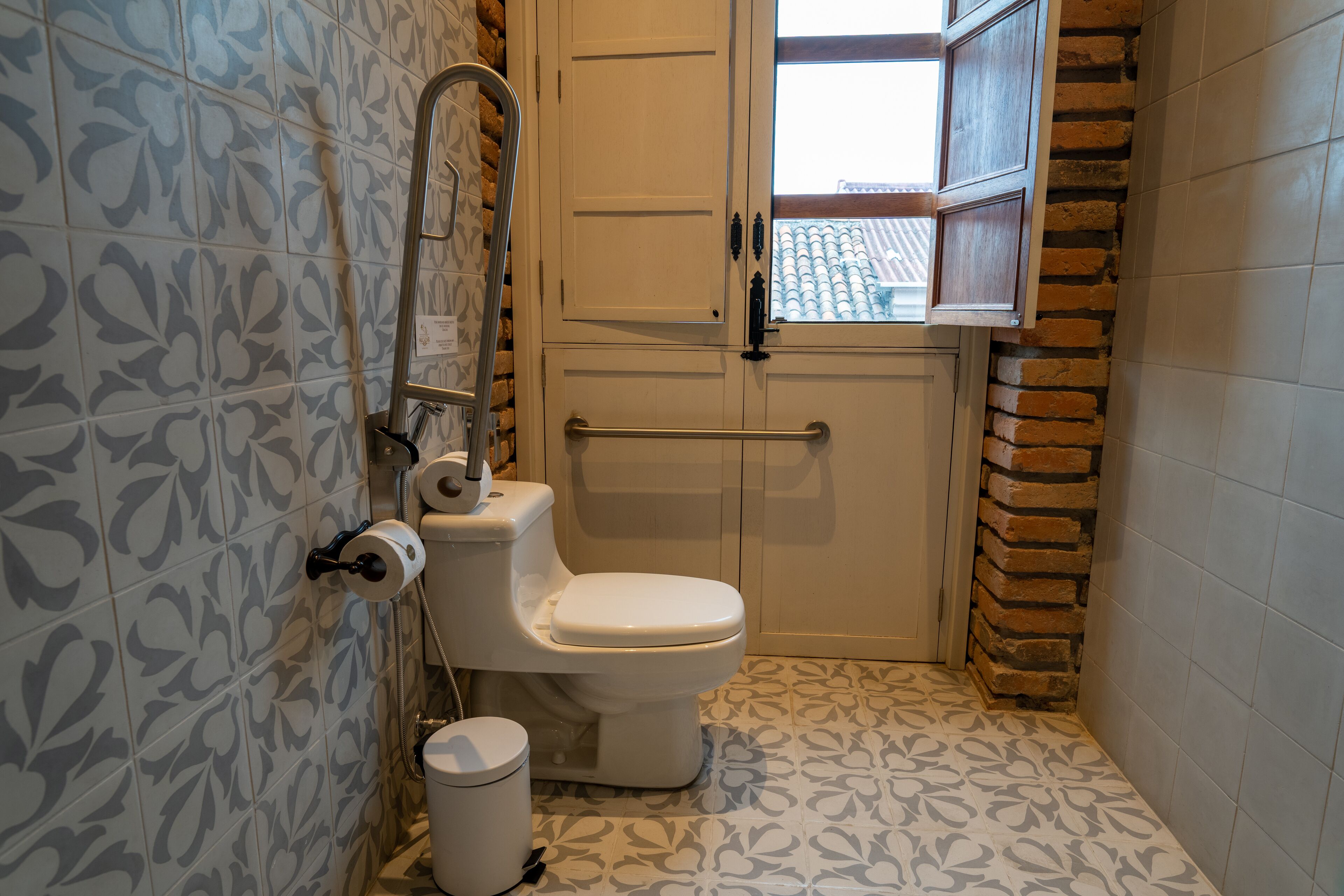 A bathroom with a toilet, a handrail, a window, and patterned tiles.