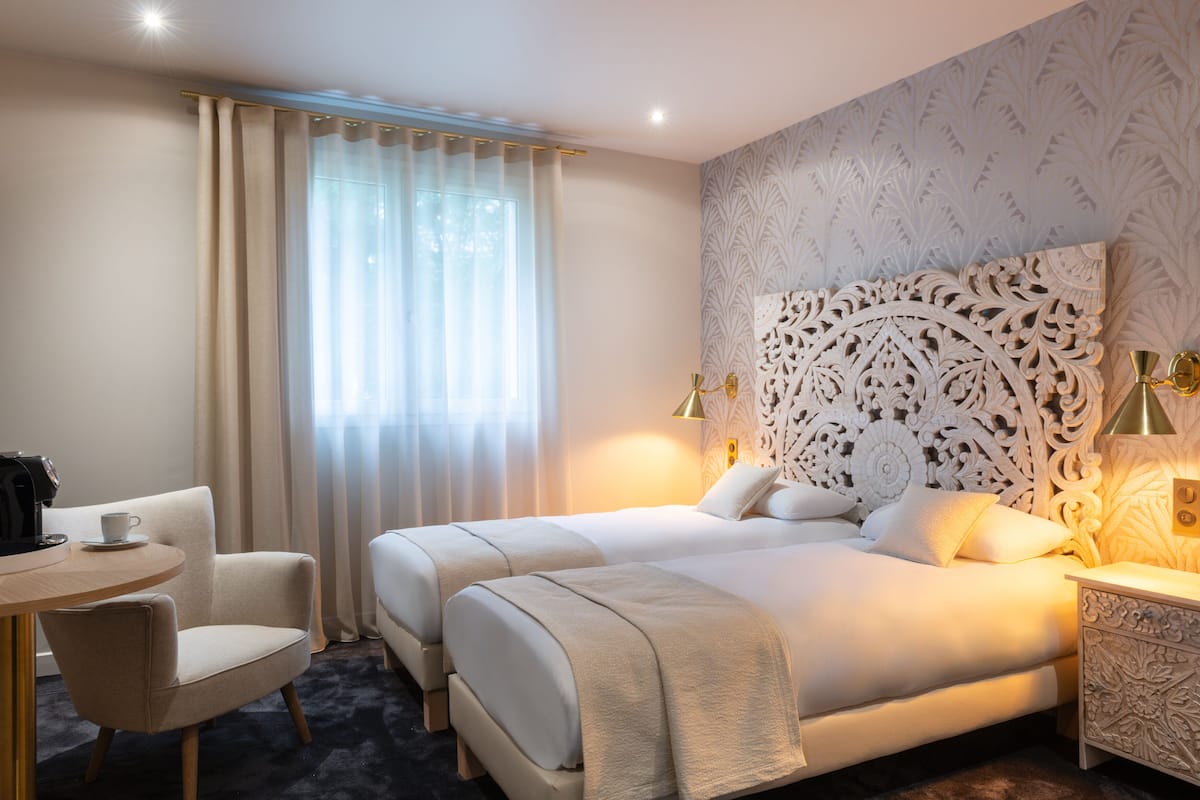 Quarto de hotel com duas camas, uma mesa redonda com uma cadeira e cabeceira decorativa.