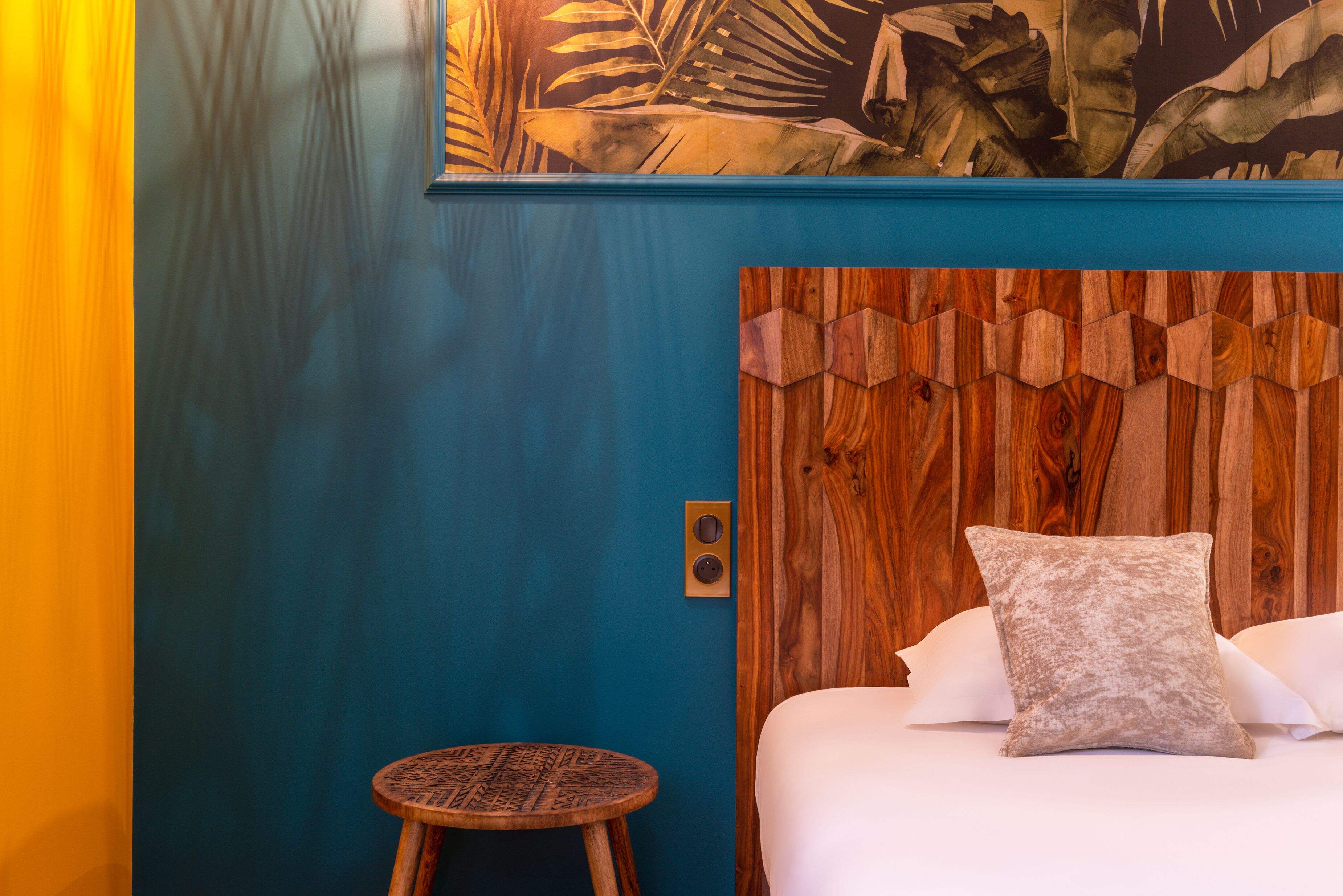 Una habitación con pared azul, cabecera de madera, cama con ropa blanca y un taburete redondo de madera.