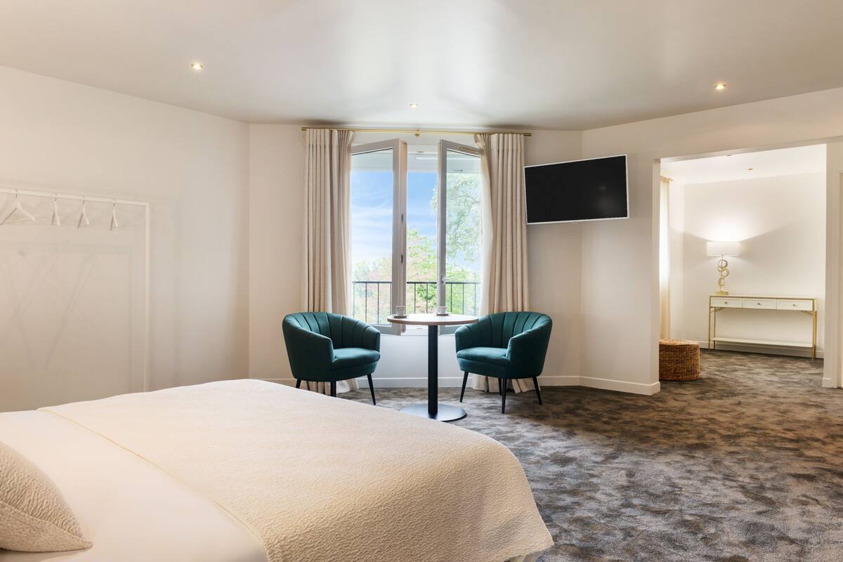 Quarto de hotel com uma cama grande, duas cadeiras, uma mesinha, televisão e janela com cortinas.