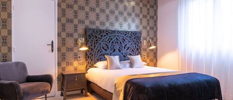 A bedroom with a patterned wallpaper, a large bed with a dark headboard, bedside lamps, a chair, and a door.