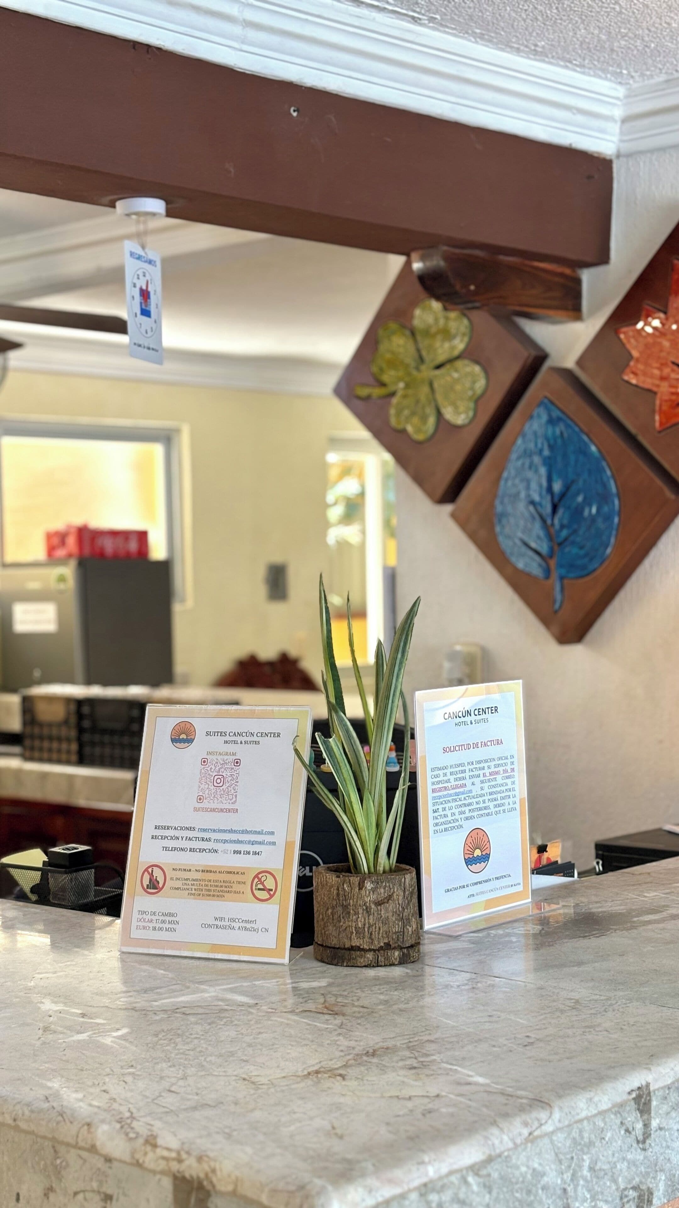 A reception desk with a plant, two informational signs, and a wall with decorative elements.
