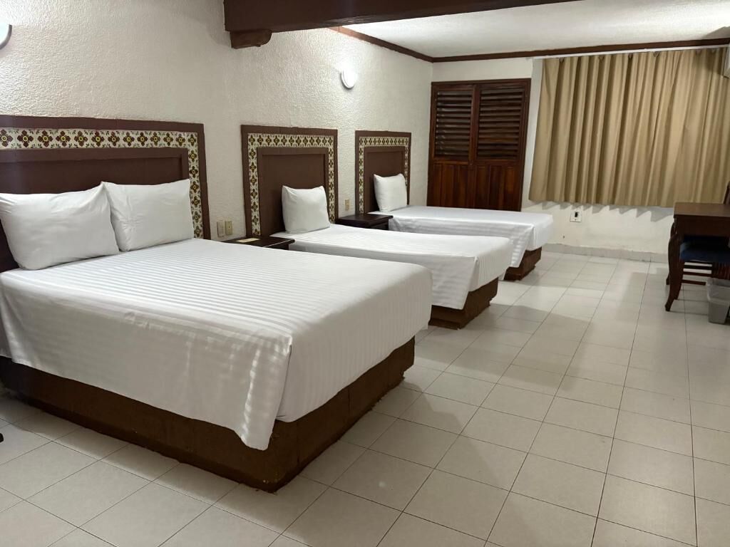 Habitación de hotel con tres camas individuales, un cabecero de madera, una mesita y una ventana con cortinas.