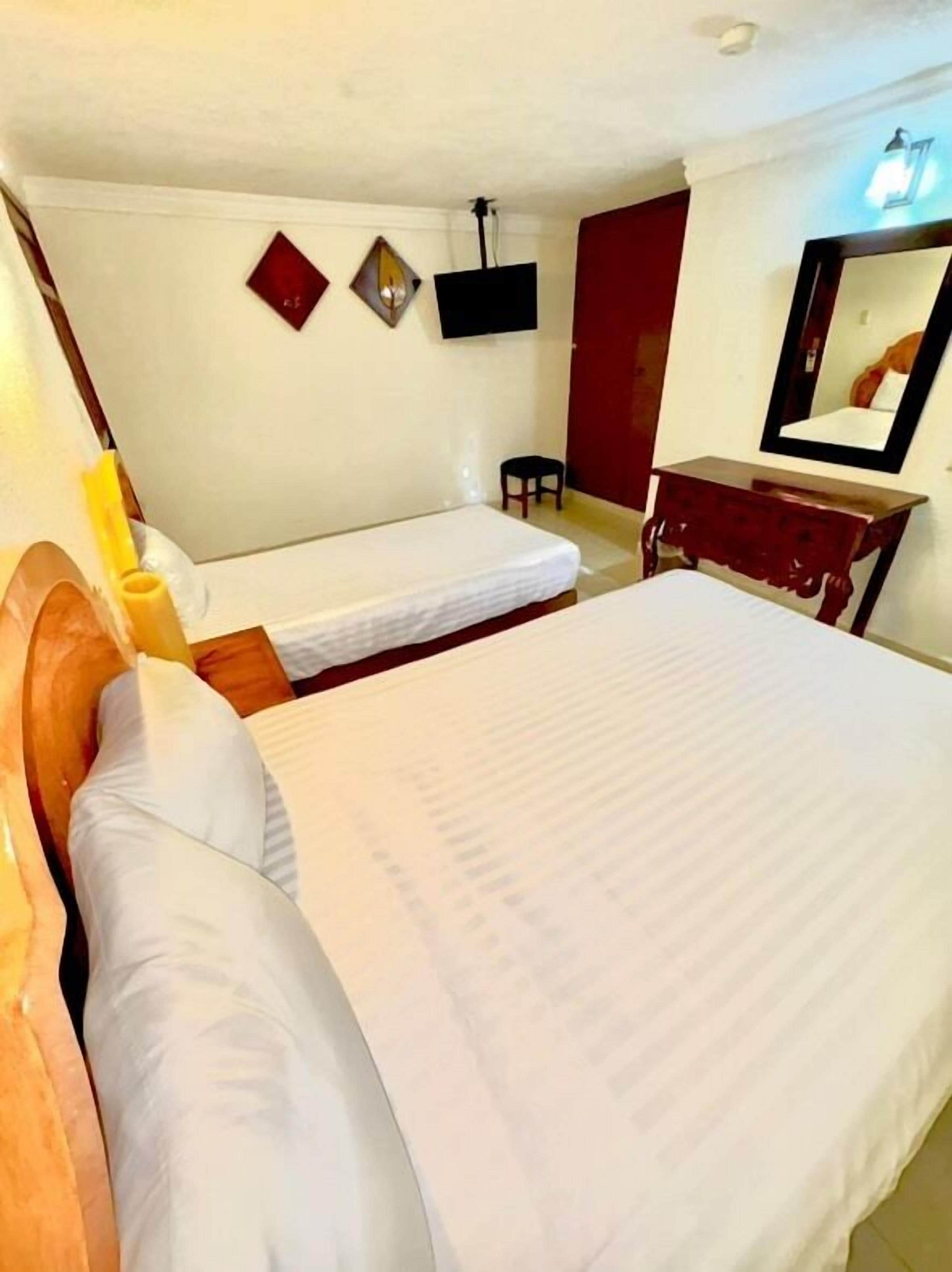 A hotel room with two single beds, a wooden headboard, a small table, a chair, a mirror, and a wall-mounted TV.