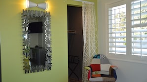 Una habitación con espejo, una silla, una ventana con persianas y una puerta que da a otra habitación.