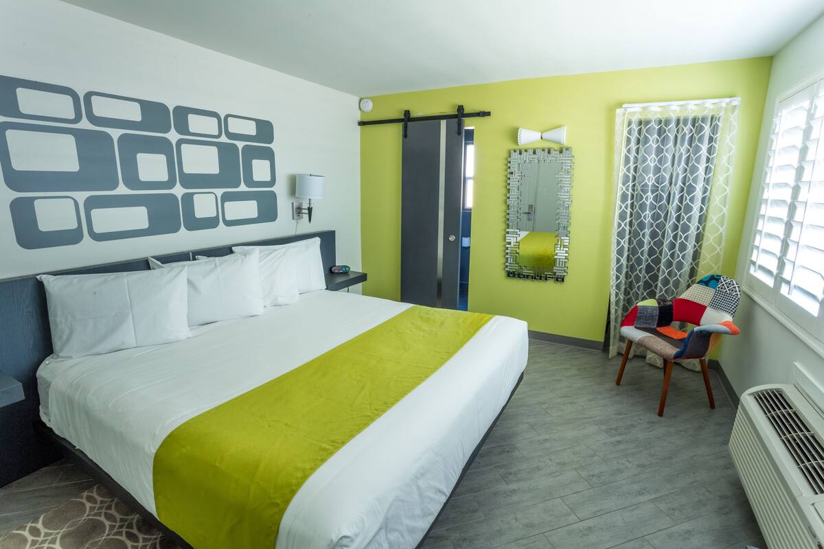 Une chambre d’hôtel avec un lit, une chaise, un miroir et une fenêtre avec des rideaux.