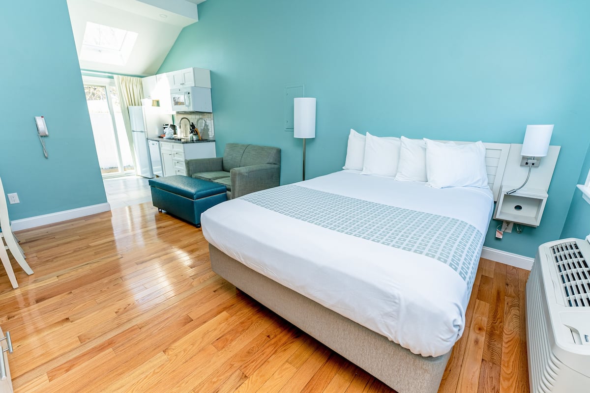 A modern hotel room with a large bed, a desk, a chair, and a kitchenette.