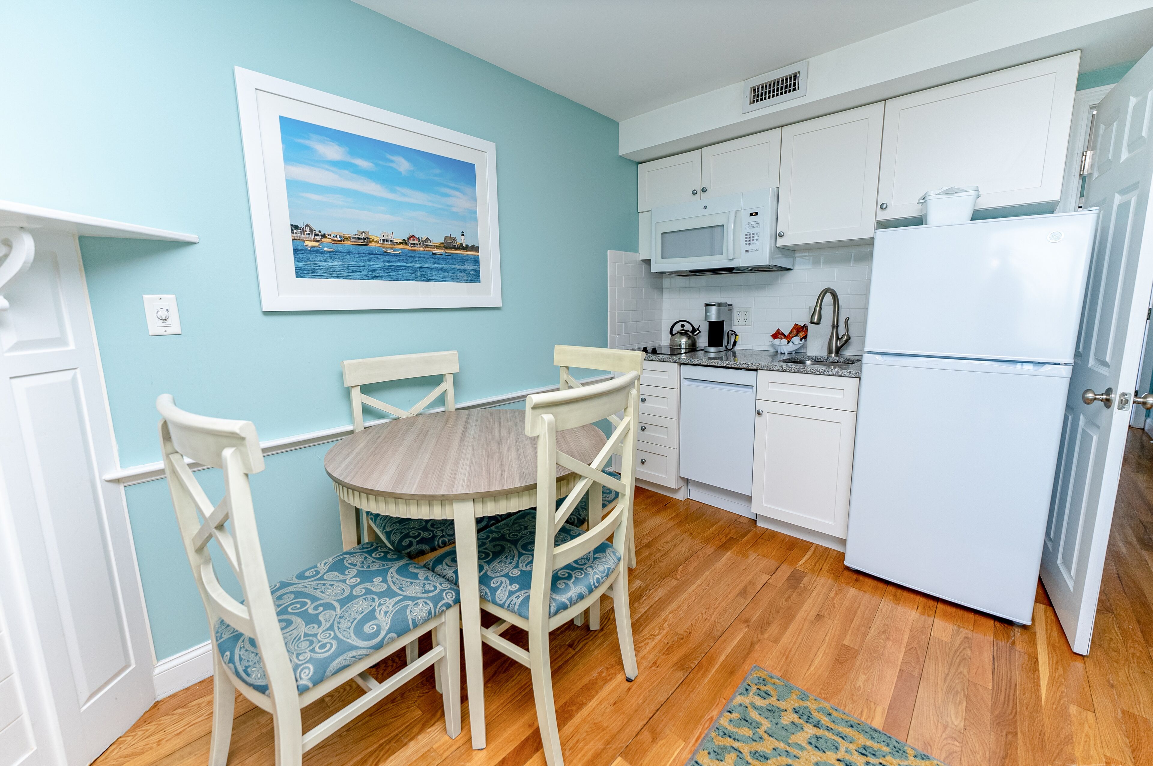A compact kitchen with white cabinets, a microwave, and a refrigerator. There is a dining table with chairs and a framed picture on the wall.