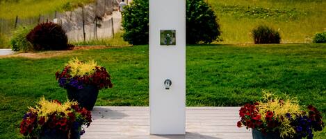 Estructura blanca con símbolo de ducha y placa, rodeada de plantas en macetas sobre una plataforma de madera, frente a un campo de césped y vistas al océano.