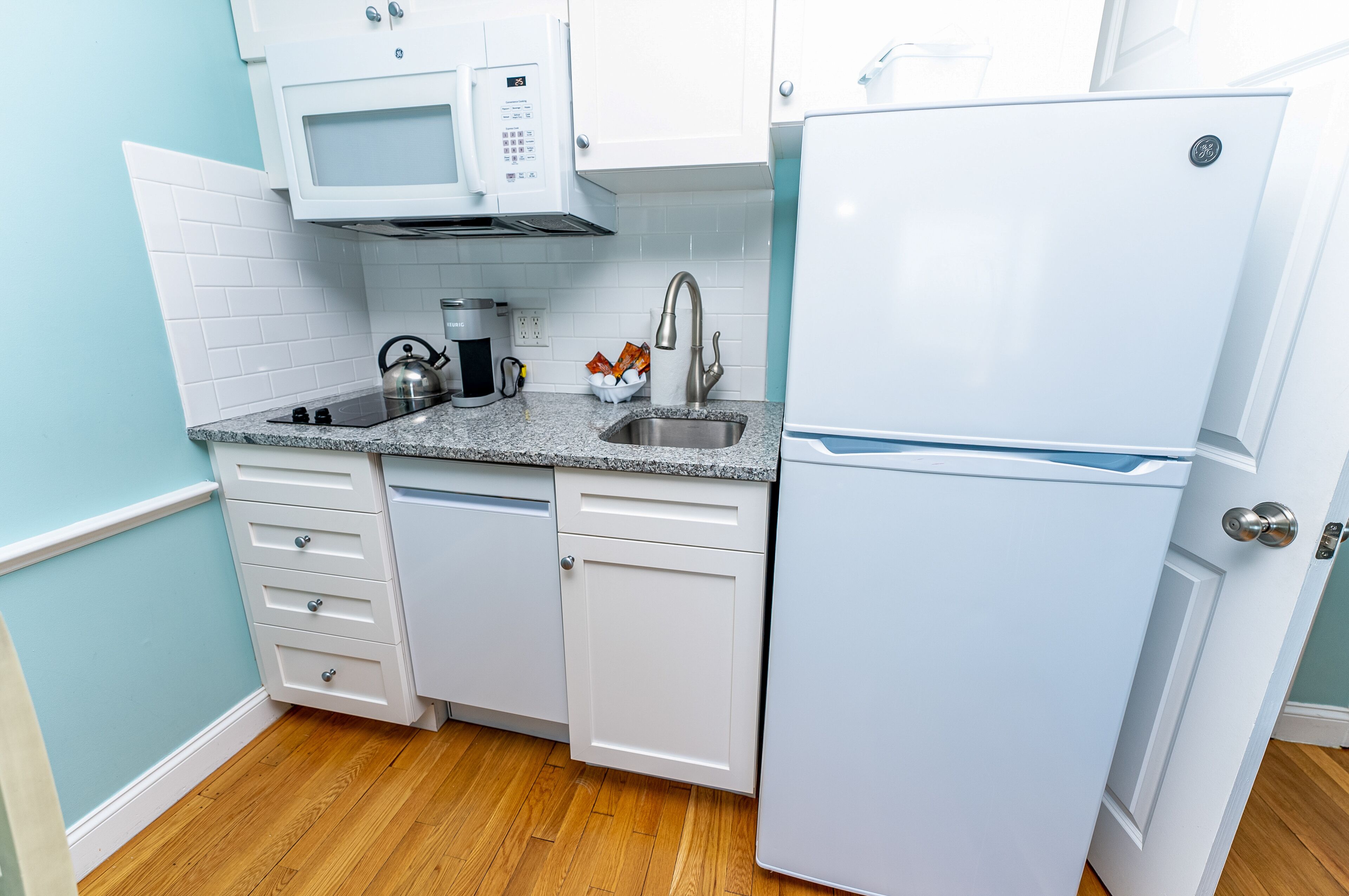 A compact kitchen with white cabinets, a built-in microwave, a stainless steel sink, and a white refrigerator.