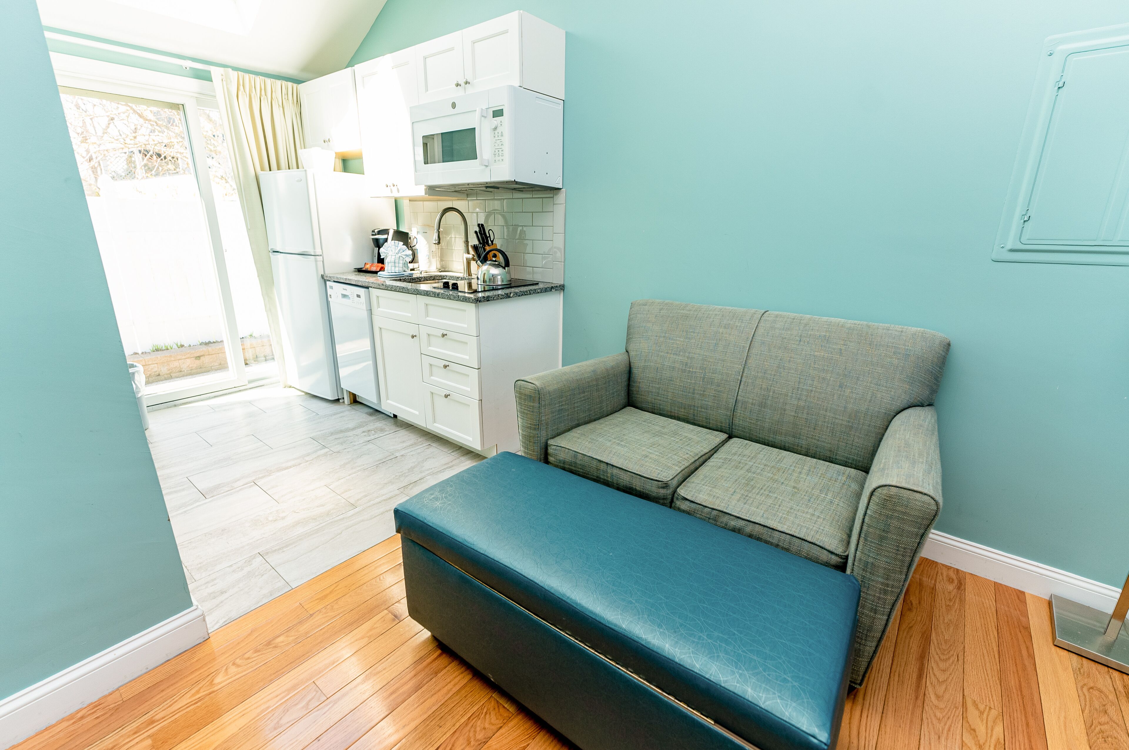 A compact kitchen with white cabinets and a microwave, a grey sofa with a blue ottoman, and a wooden floor.