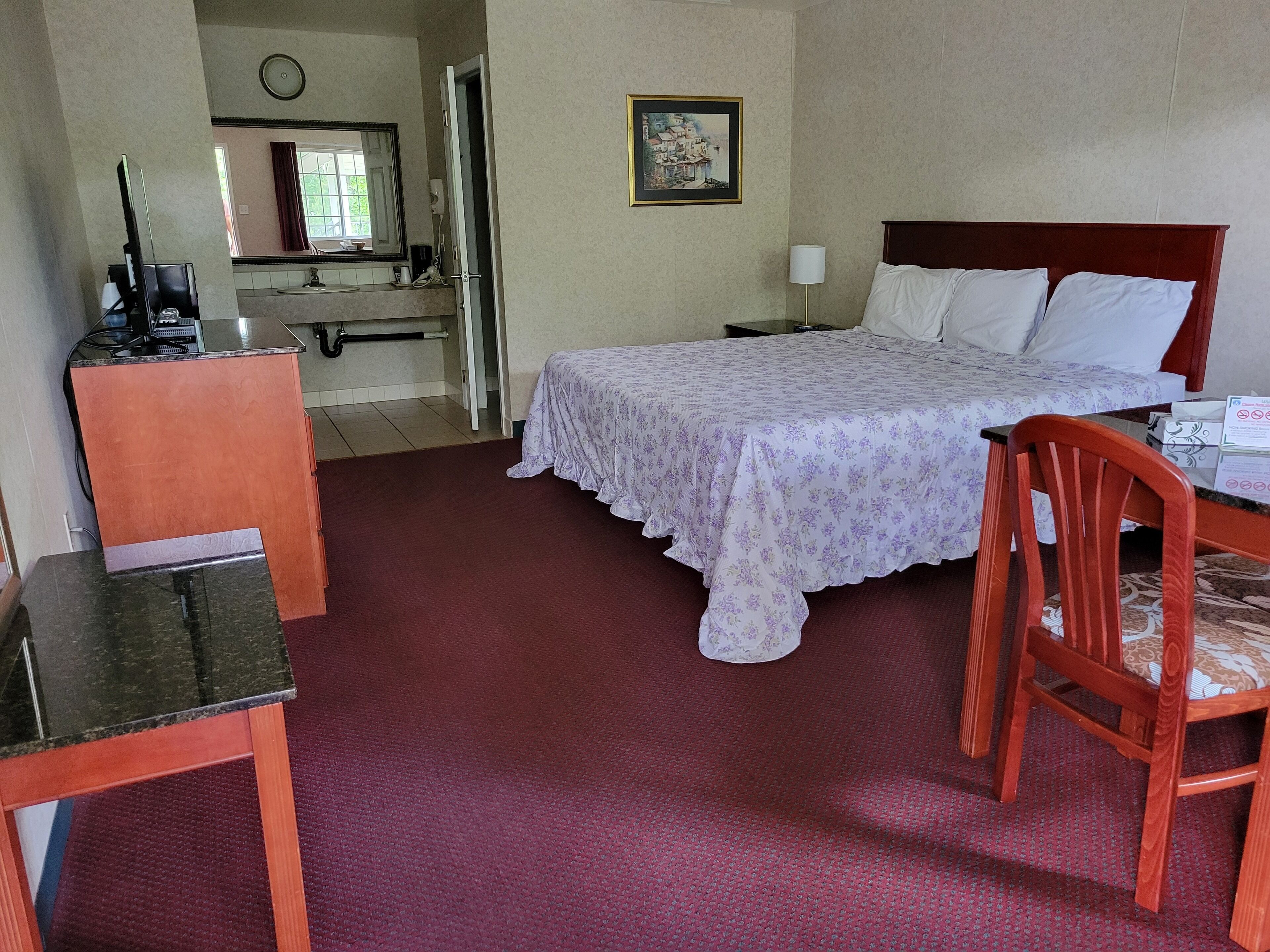 A hotel room with a bed, a desk, a chair, a mirror, a picture on the wall, and a clock.
