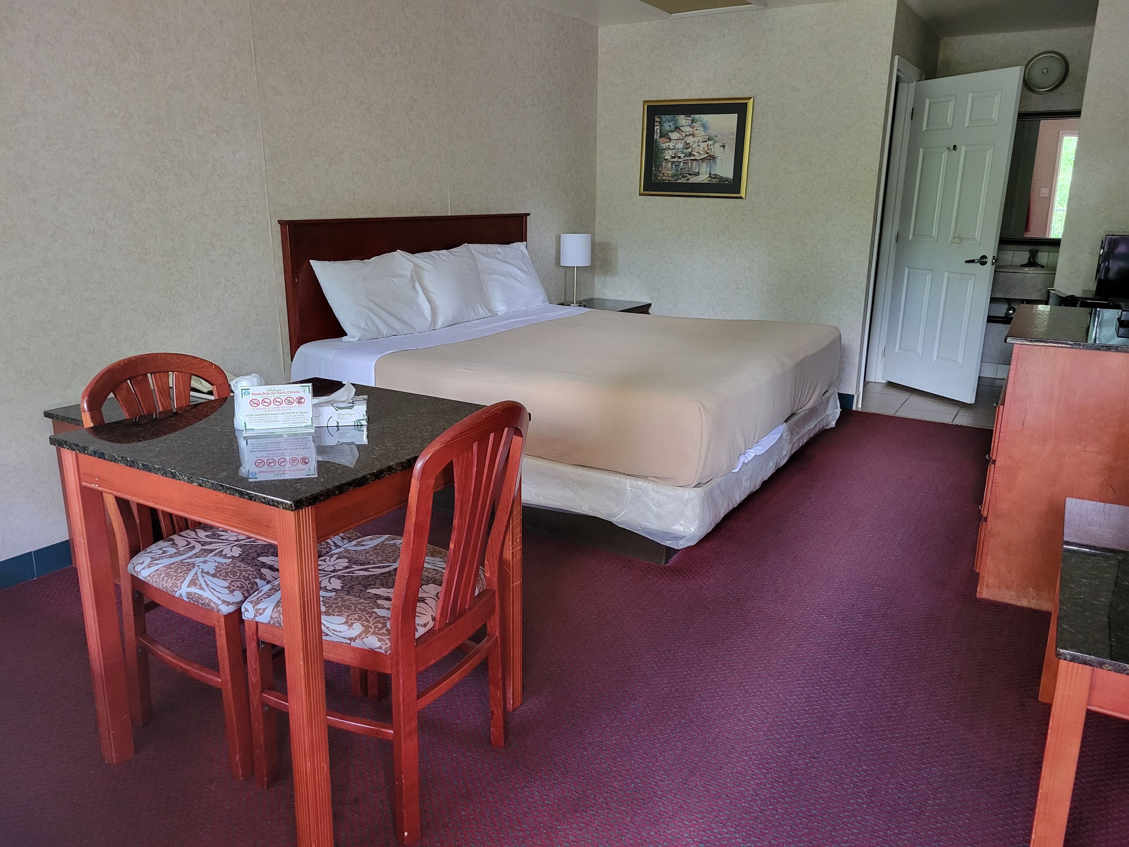 A hotel room with a bed, a desk with chairs, a framed picture on the wall, and a door leading to the kitchen.