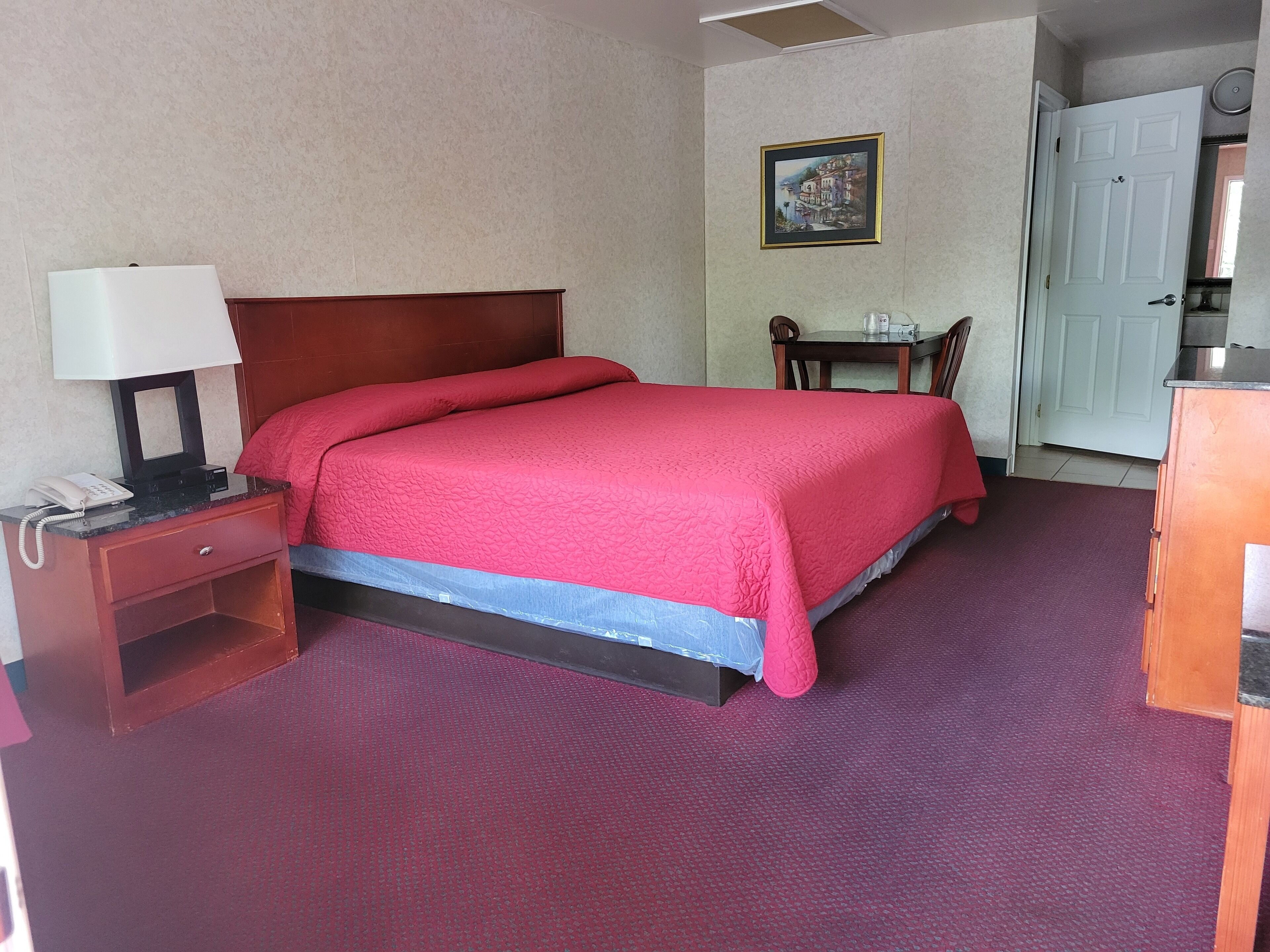 Una camera d'albergo con un letto, un comodino con una lampada, un tavolino con una sedia e una porta.