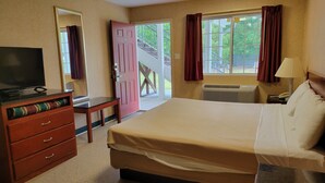 A hotel room with a bed, a desk with a television, a dresser, a window with curtains, and a view of the outdoors.