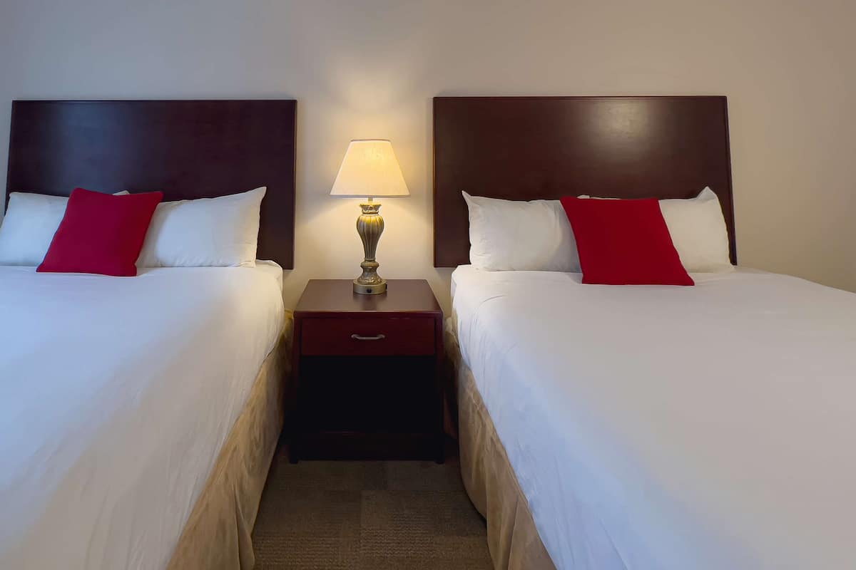 Habitación de hotel con dos camas, cada una con ropa blanca y almohadas rojas, una mesita de noche con una lámpara y un cabecero de madera.