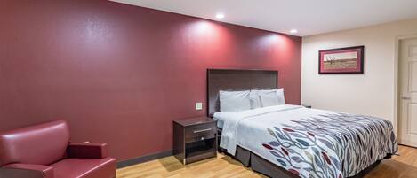 A hotel room with a bed, a nightstand, a red chair, and a wall-mounted picture.