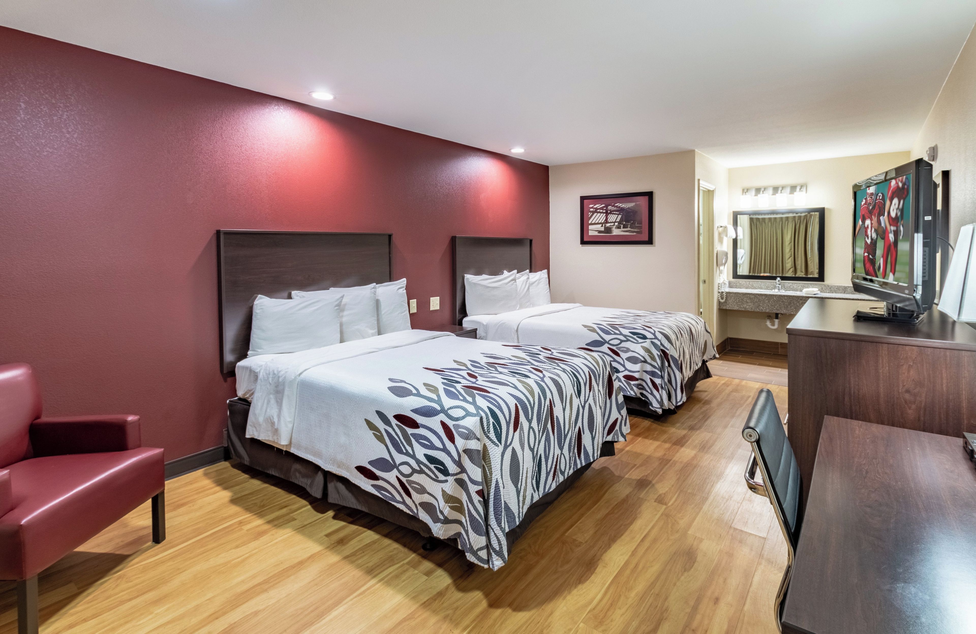 Habitación de hotel con dos camas, un escritorio, una silla, un televisor y un baño con espejo y lavamanos.