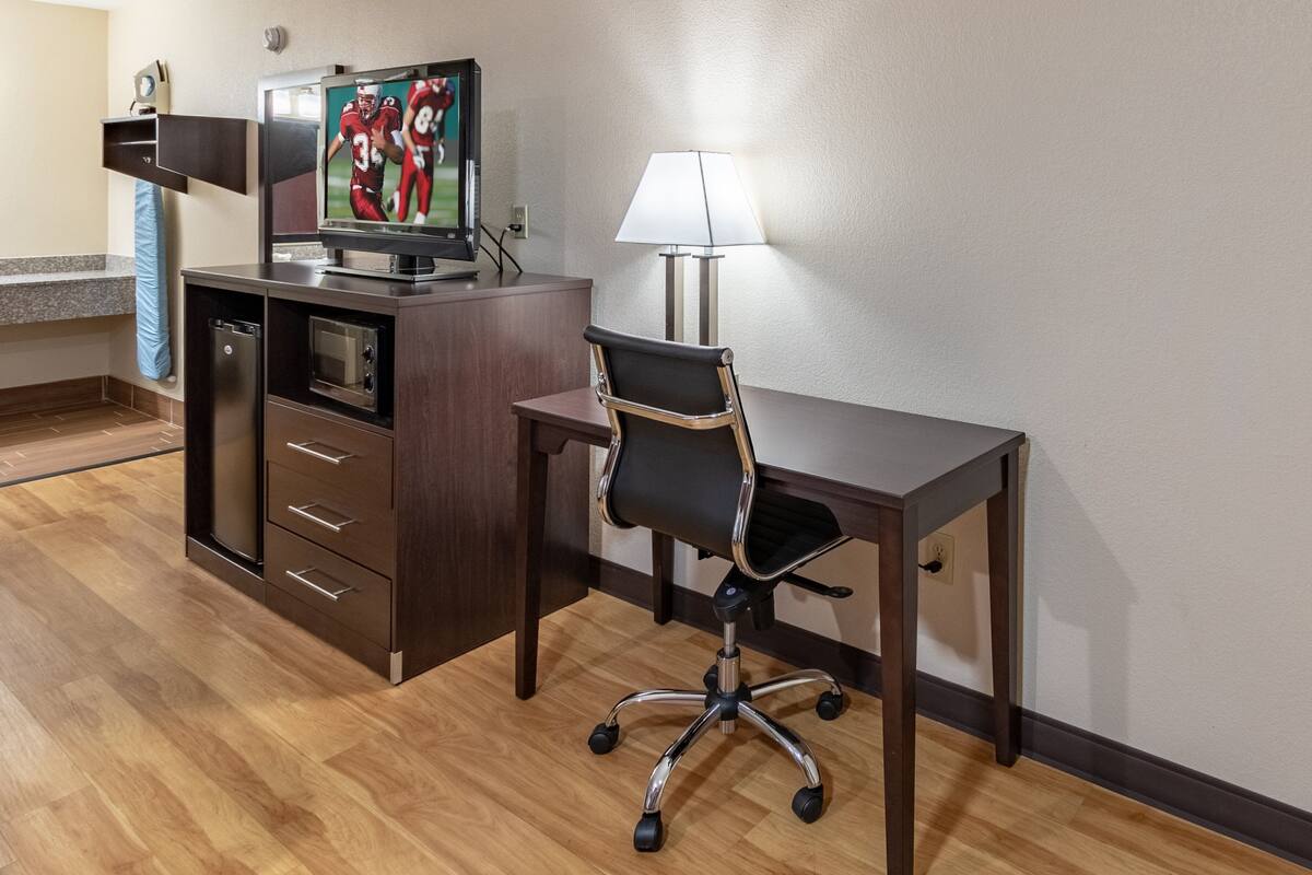 Ein Hotelzimmer mit einem Holztisch, einem schwarzen Bürostuhl, einem Fernseher auf einem Schrank und einer Lampe auf dem Tisch.