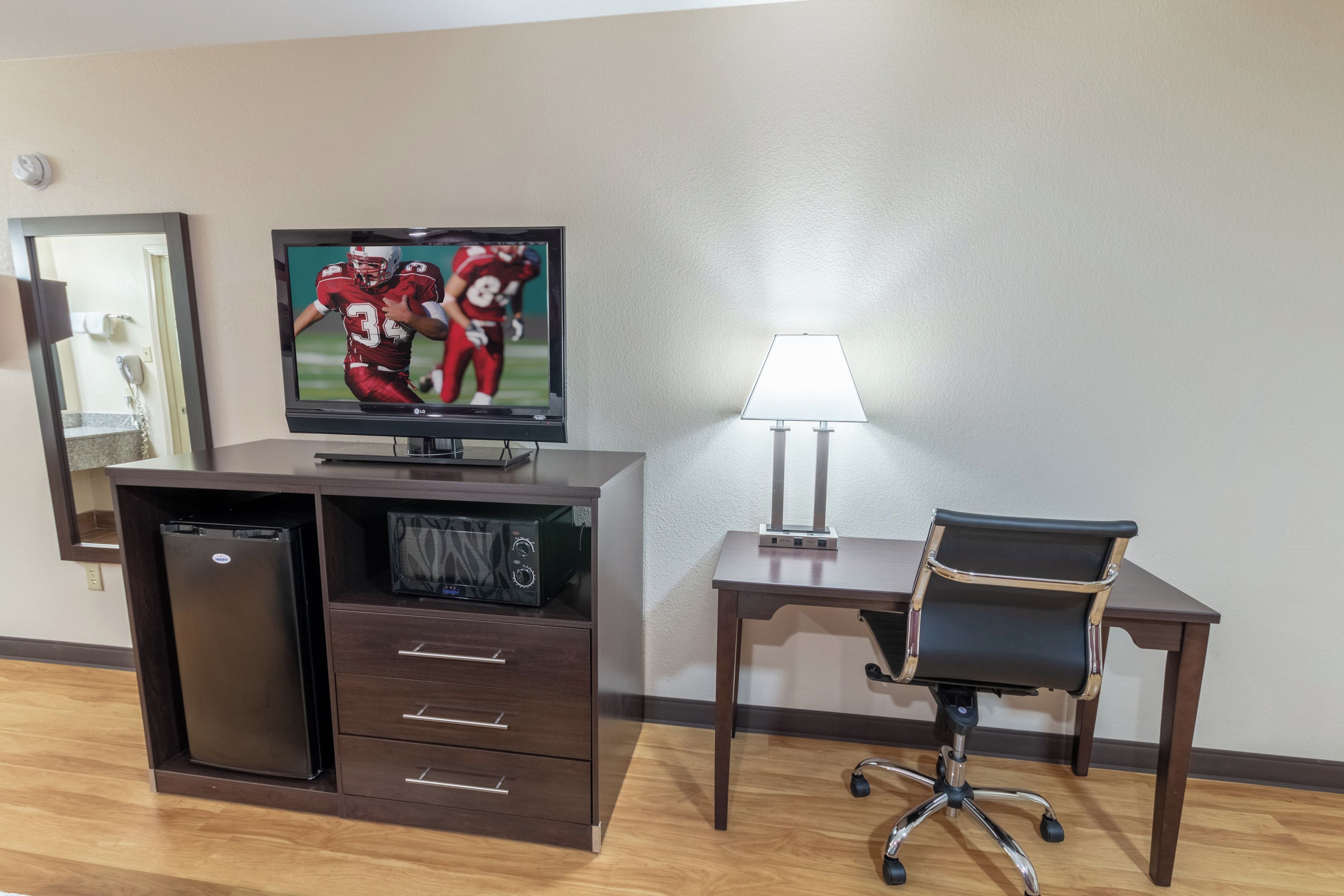 A hotel room with a TV, a desk with a lamp, a chair, a mini-fridge, and a built-in microwave.