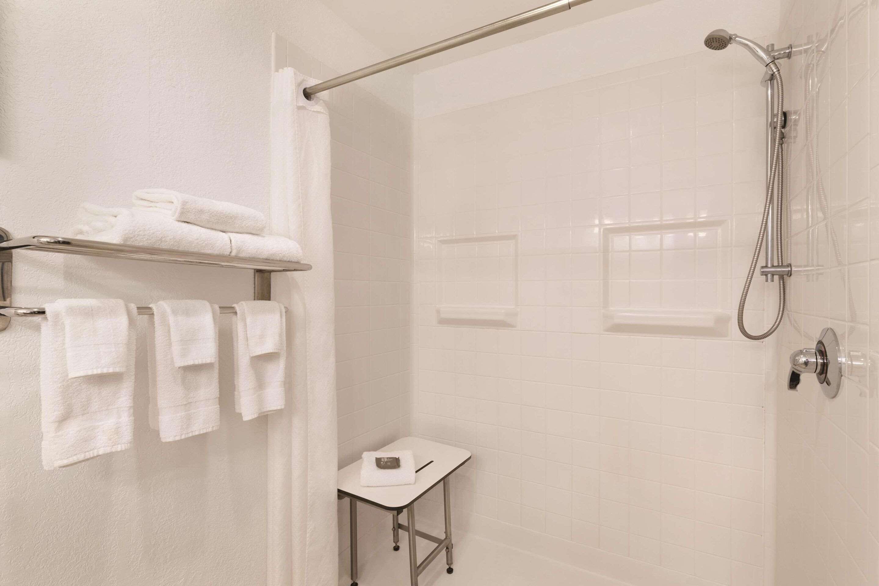 A bathroom with white tiled walls, a shower with a handheld showerhead, a towel rack with folded towels, and a small table with a towel.