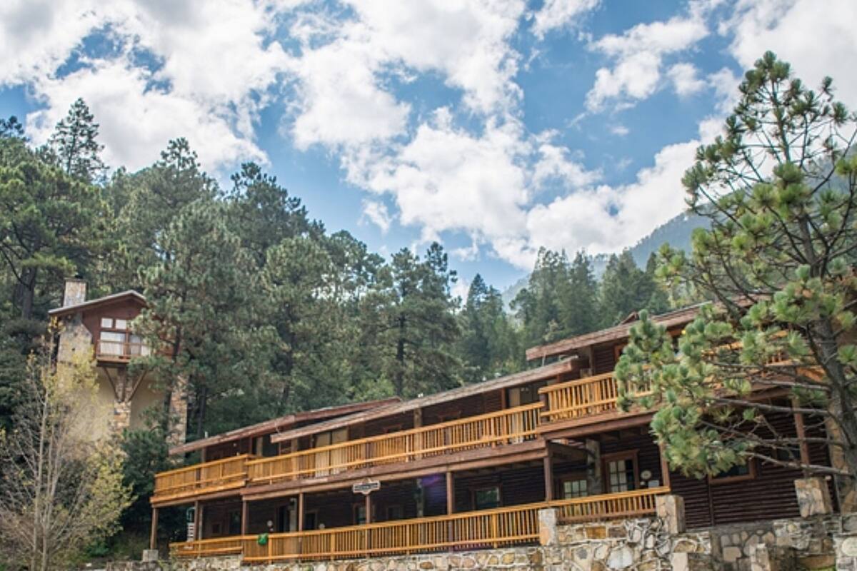 A rustic lodge with a stone tower and wooden balconies, surrounded by dense forest and mountains.