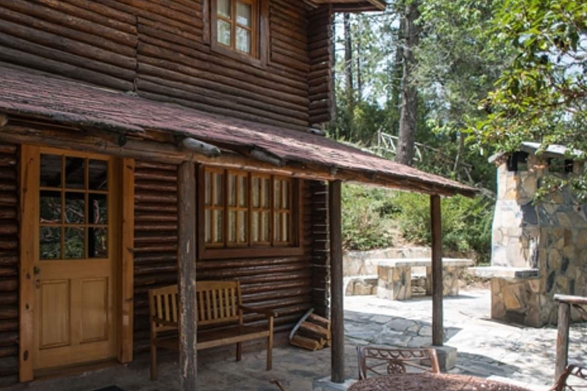 A rustic log cabin with a wooden door and windows, a porch with benches, and a stone wall in the background.