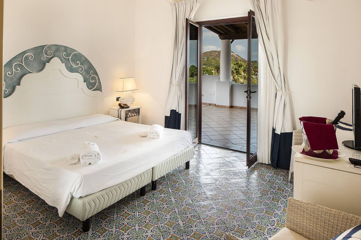 A hotel room with a bed, a nightstand, a lamp, a red chair, and a view of the outdoors.