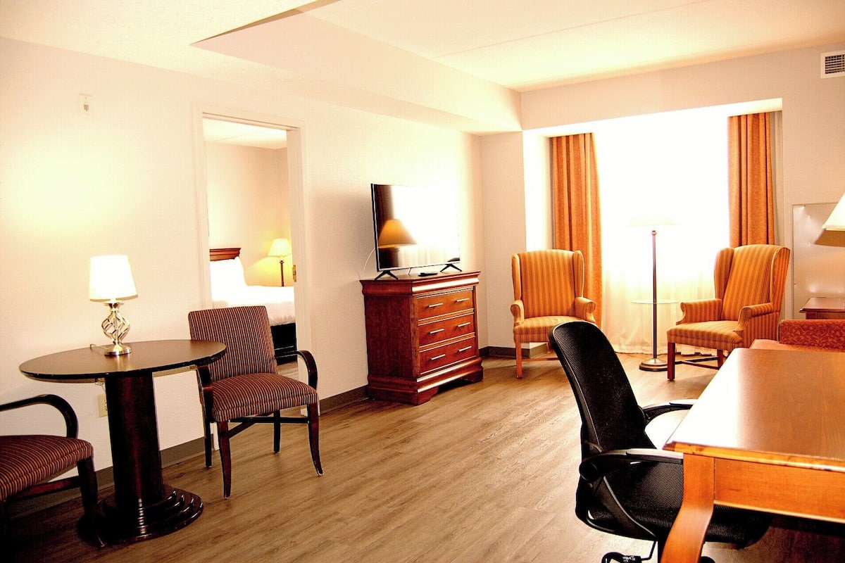 A hotel room with a wooden floor, a desk with a lamp, two striped armchairs, a sofa, a TV on a wooden cabinet, and a large window with curtains.