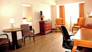A hotel room with a wooden floor, a desk with a lamp, two striped armchairs, a sofa, a TV on a wooden cabinet, and a large window with curtains.