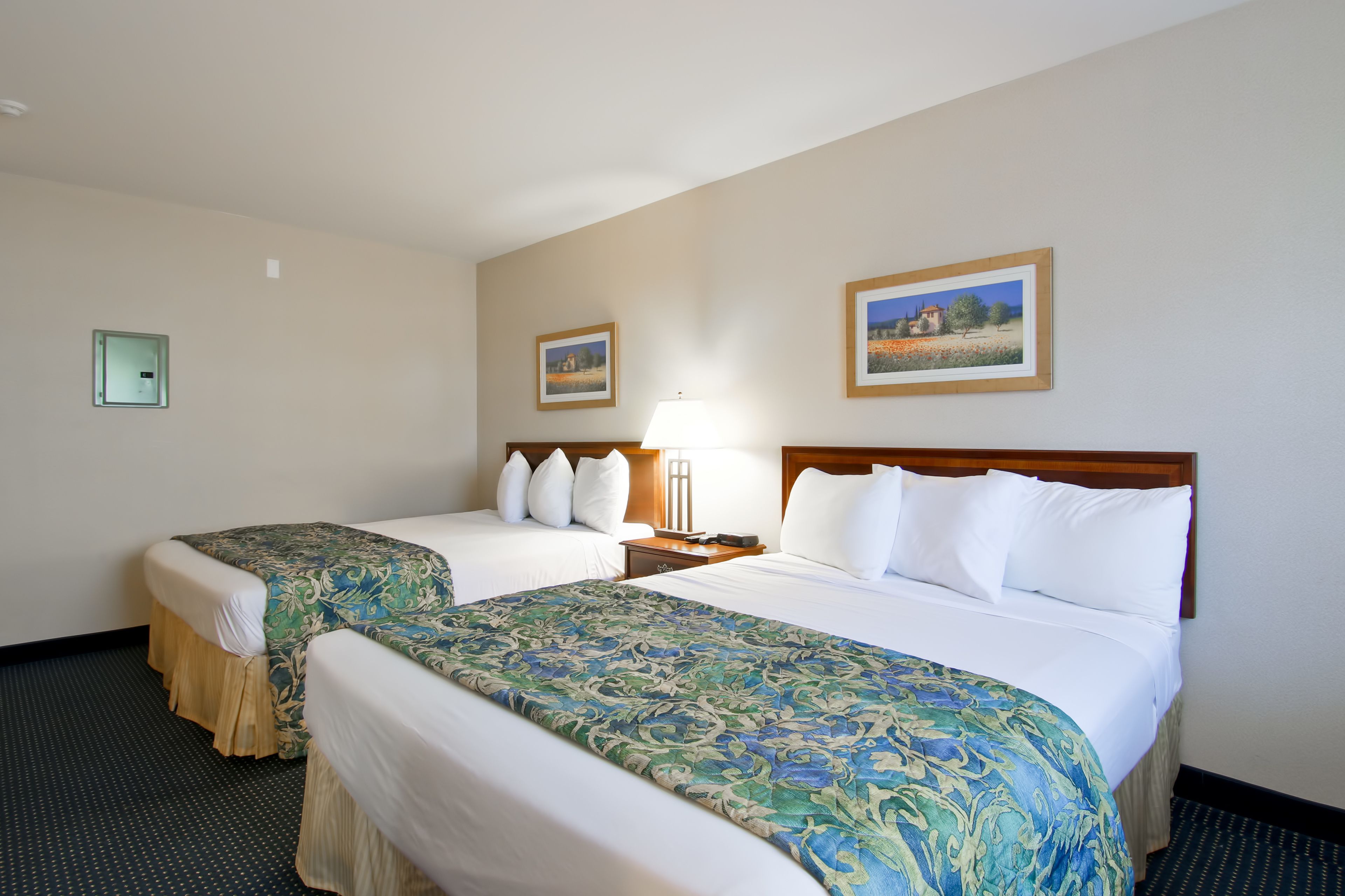 Quarto de hotel com duas camas, uma mesa de cabeceira, uma luminária e duas fotos emolduradas na parede.