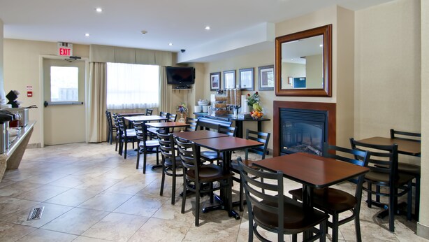 A hotel dining area with multiple tables and chairs, a television mounted on the wall, and a fireplace. - Travelodge by Wyndham Trenton (Quinte West)