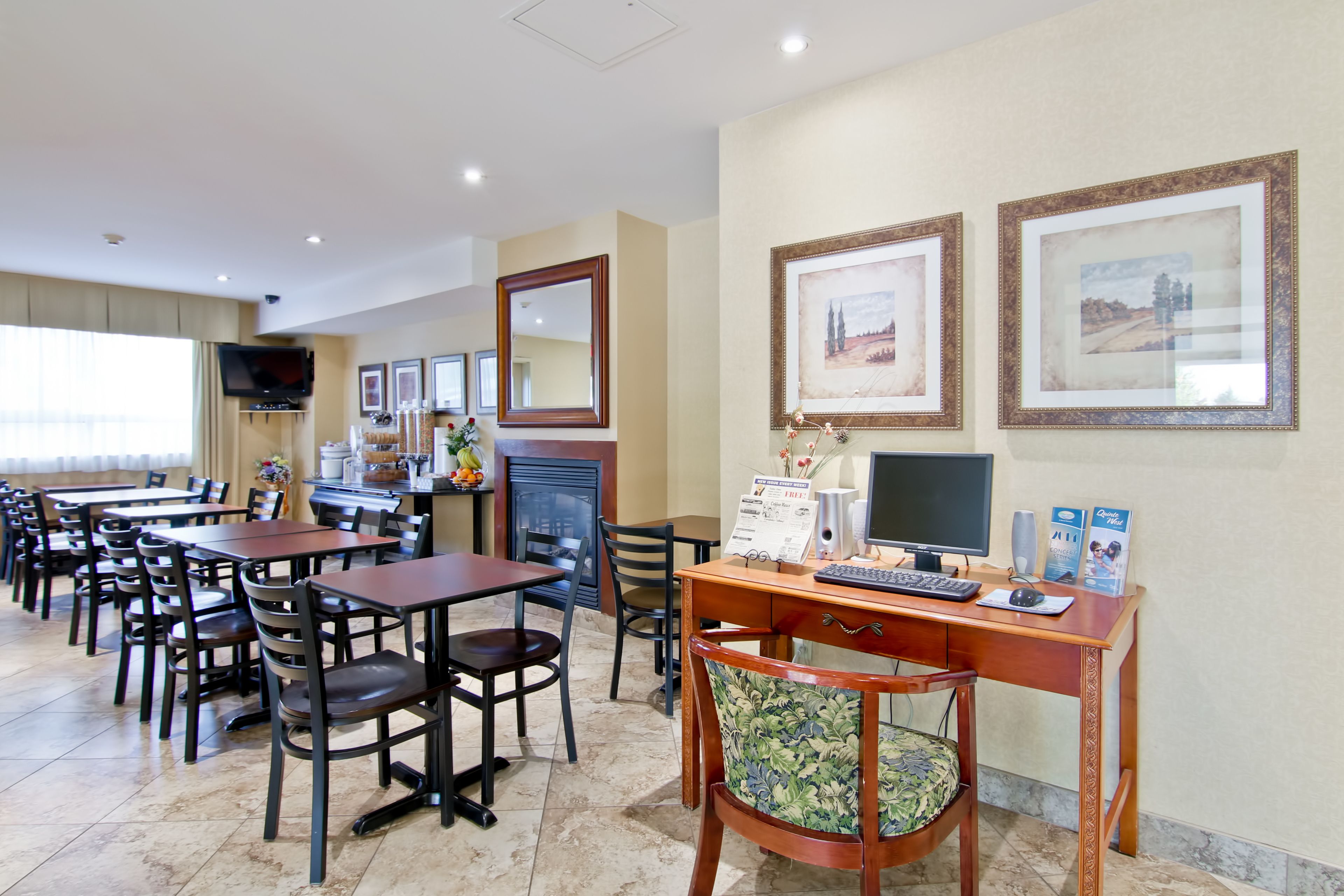A hotel dining area with multiple tables and chairs, a fireplace, a desk with a computer, and framed pictures on the wall.