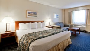 A hotel room with a large bed, two bedside tables with lamps, a sofa, a small table, and a window with curtains. - Travelodge by Wyndham Trenton (Quinte West)