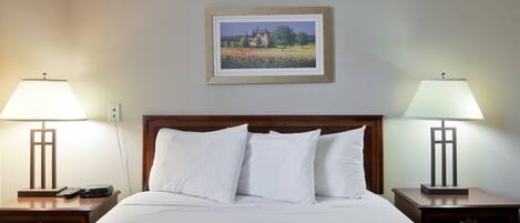 A hotel room with a large bed, two bedside tables with lamps, a framed picture on the wall, and a wooden headboard.