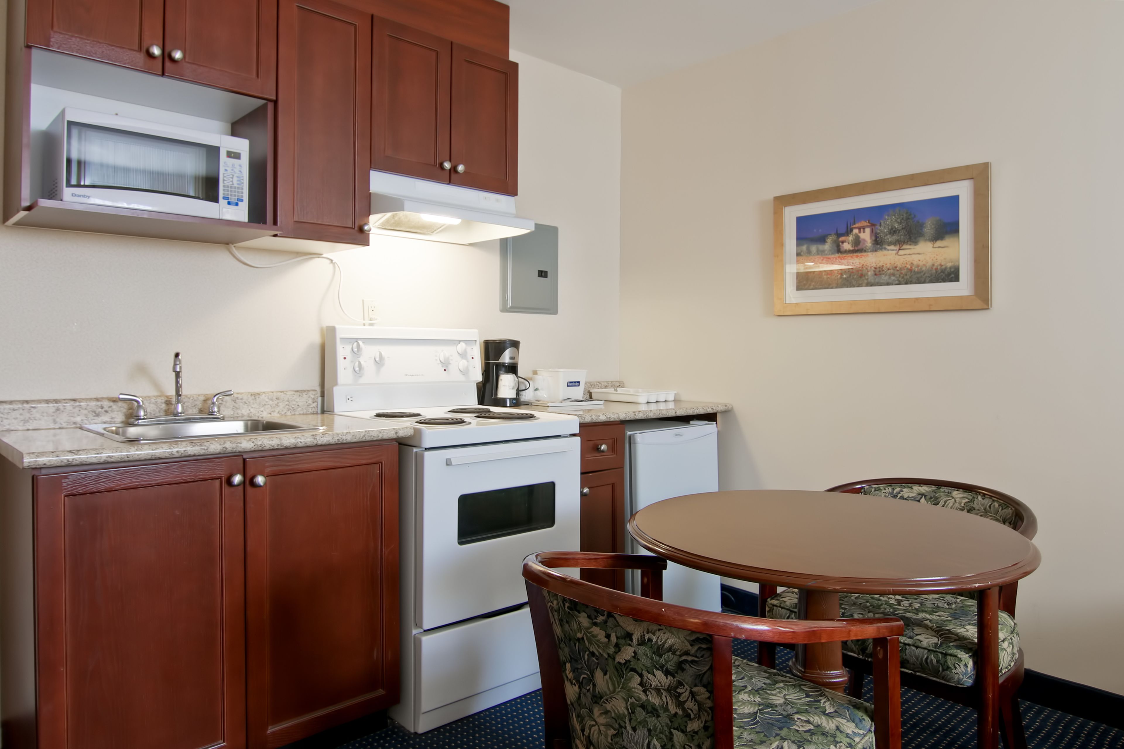 A compact kitchen with a microwave, stove, sink, and a small refrigerator. There is a dining table with two chairs and a framed picture on the wall.