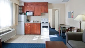 A compact kitchen with wooden cabinets, a microwave, a refrigerator, and a stove. There is a dining table with a chair and a sofa in the living area. - Travelodge by Wyndham Trenton (Quinte West)