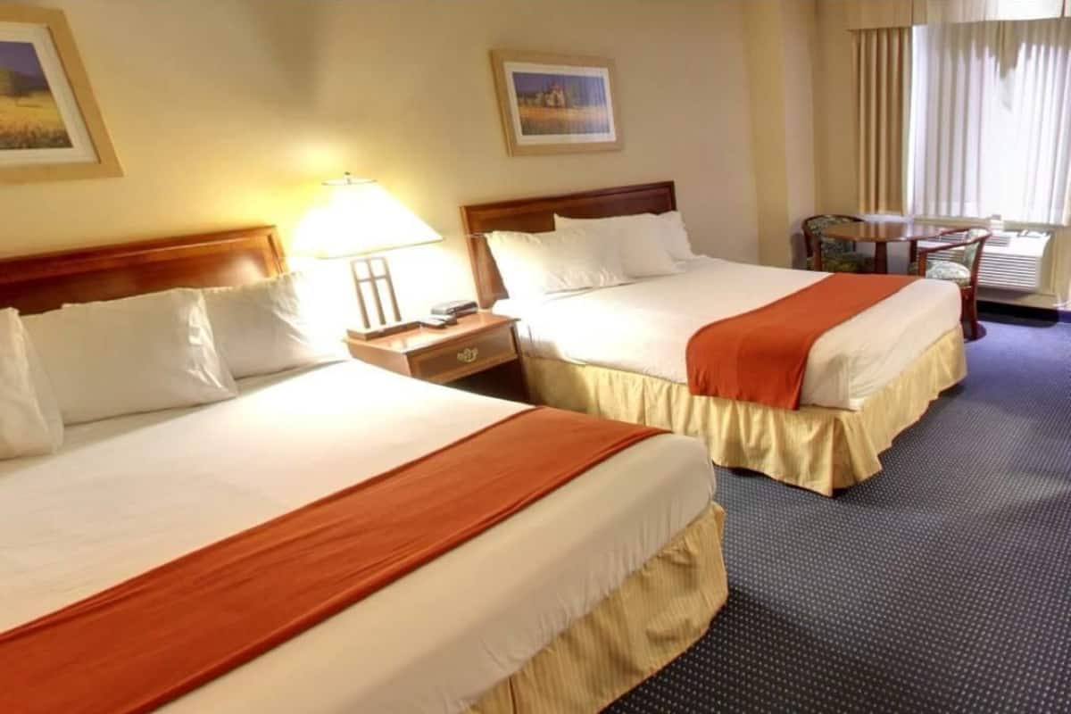 A hotel room with two beds, a nightstand, a lamp, a chair, and a window with curtains.