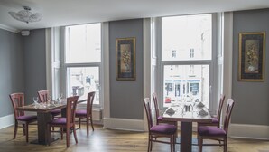 Un espace repas avec une table et des chaises en bois, de grandes fenêtres et des œuvres d’art encadrées accrochées aux murs.