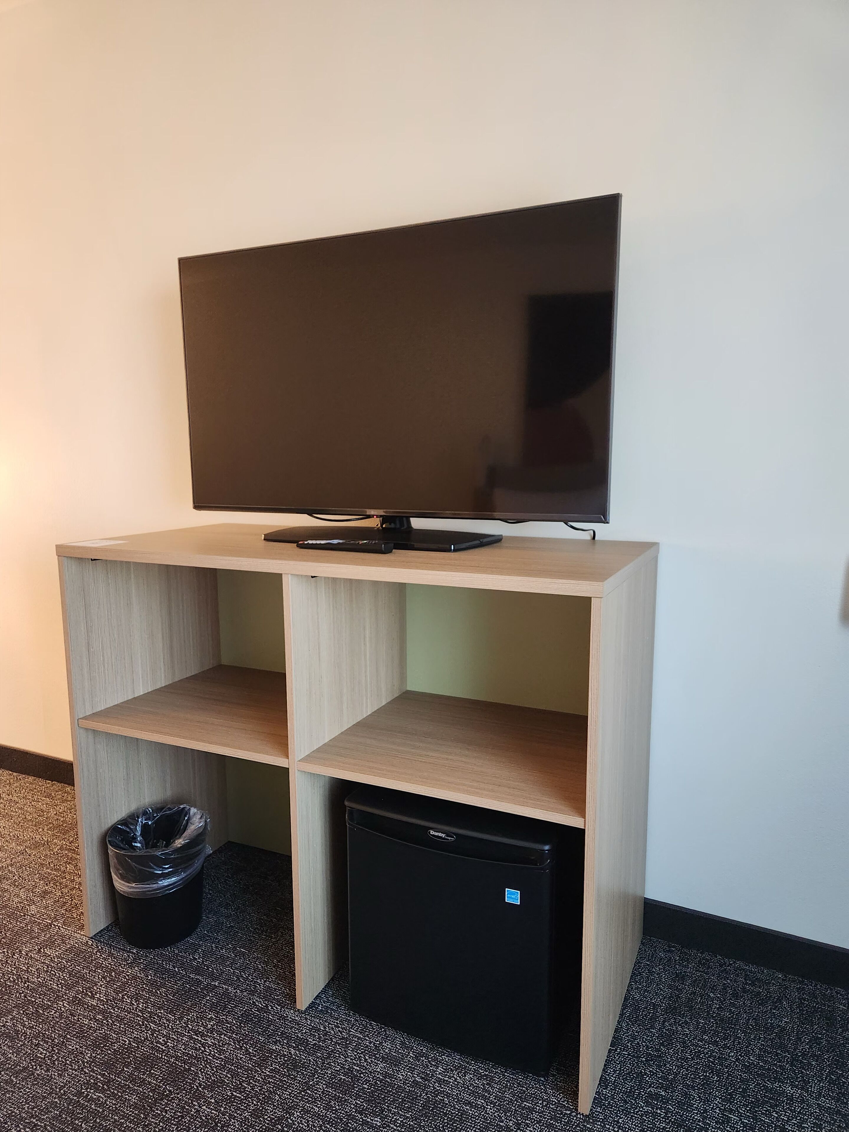 Un mueble de televisión de madera con un televisor de pantalla plana, un refrigerador negro pequeño y un cesto de basura.